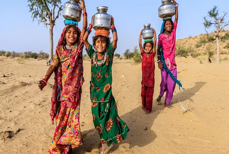 Girls carrying water pots on their heads across a desert village